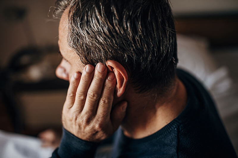A man with short hair is sitting and touching his left ear, possibly due to an ear blockage or listening carefully. He is wearing a dark sweater, and the blurred background hints at an indoor setting.