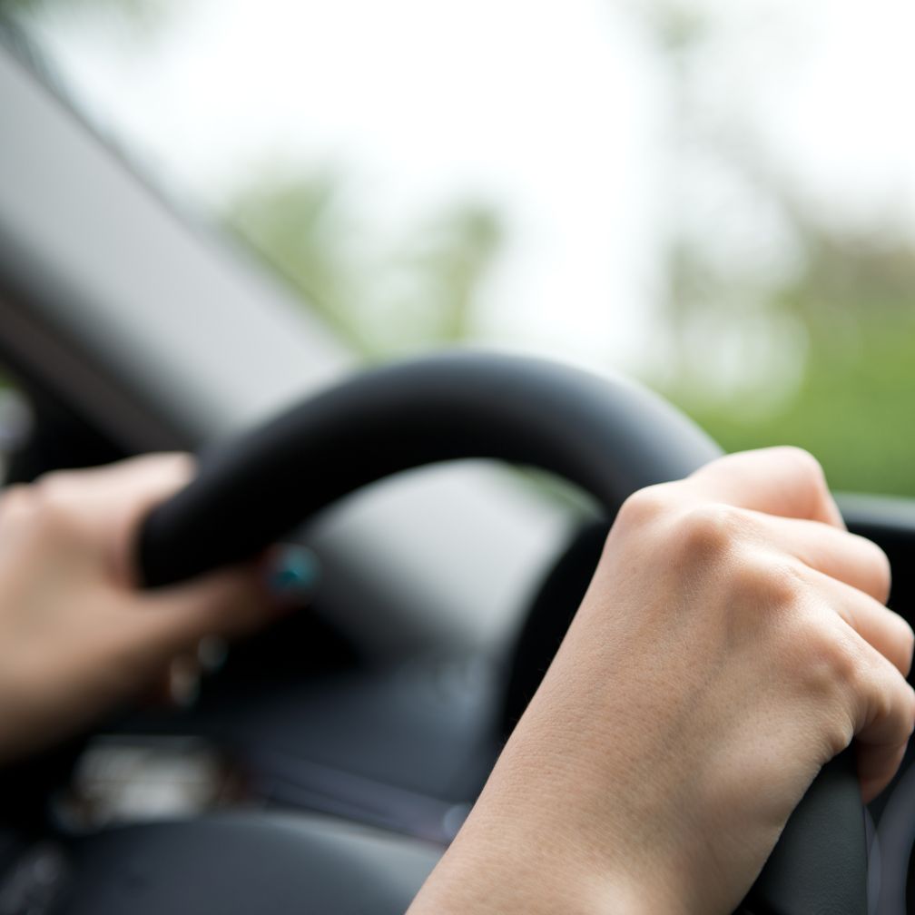 Close-up of a persons hands gripping a steering wheel while driving a car, with a blurred view of the car interior and greenery outside the window in the background.