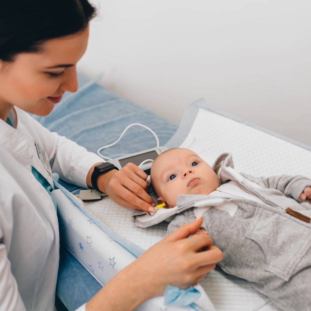 A healthcare professional performs a hearing test on a baby lying on an exam table, using specialized equipment. The baby looks up while the professional gently holds a device to the babys ear.