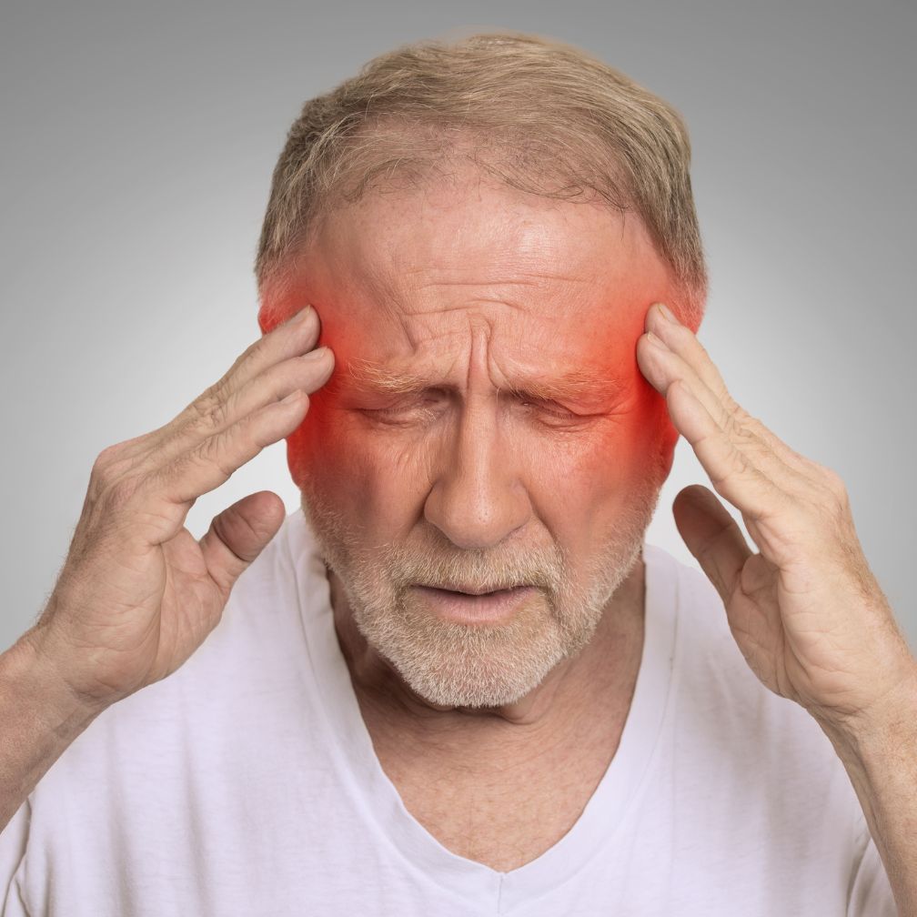 Older man with gray hair touching his temples, eyes closed, appearing to be in pain. Red coloring is shown on his temples, suggesting a headache or migraine. He is wearing a white shirt and standing against a gray background.