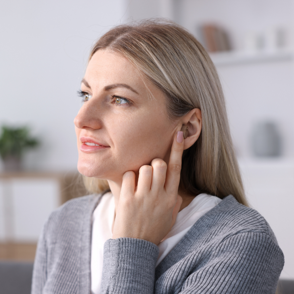 A woman with blonde hair touches her ear with one finger, appearing to listen carefully. She is wearing a gray sweater and is indoors, with blurred shelves and plants in the background.