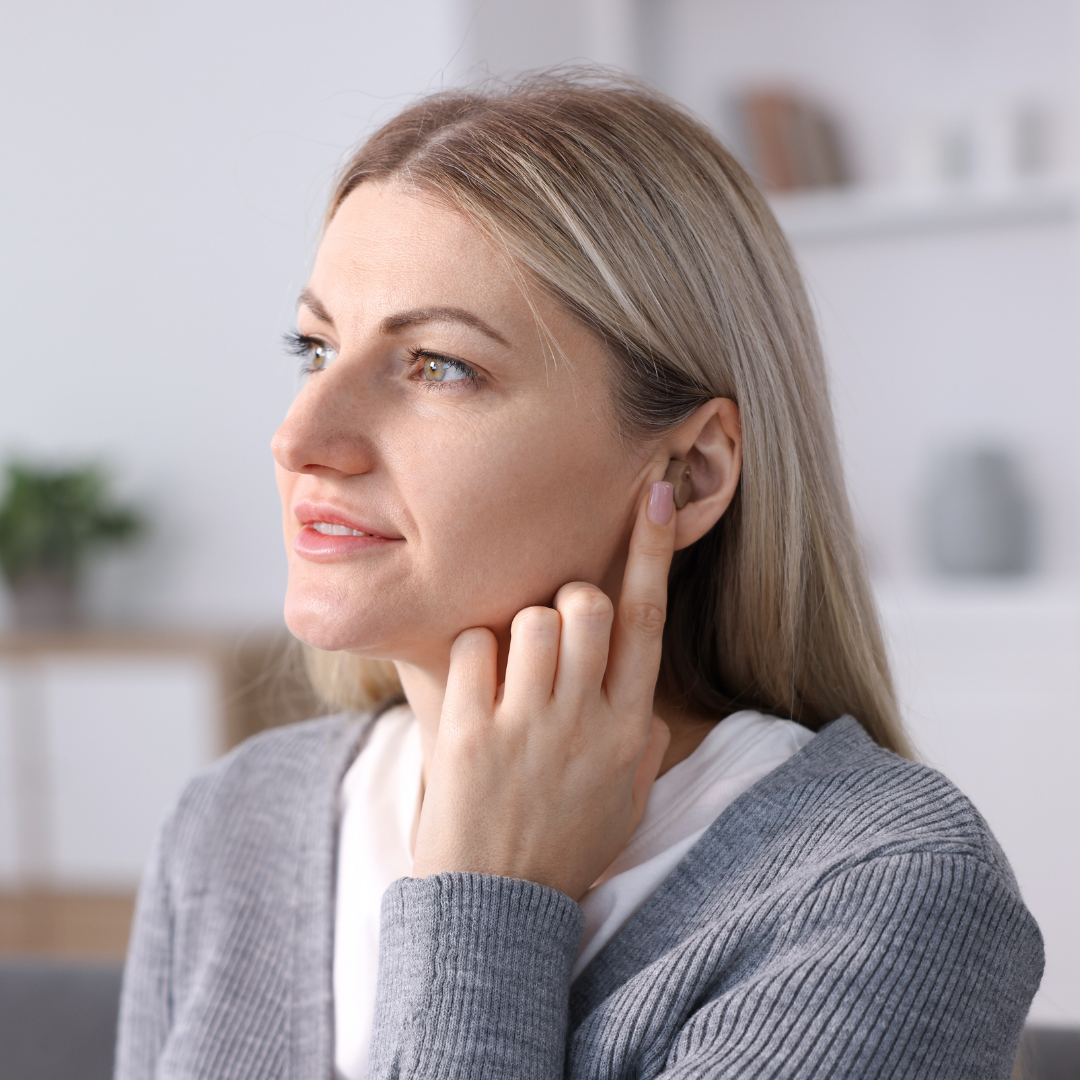 A woman with blonde hair touches her ear with one finger, appearing to listen carefully. She is wearing a gray sweater and is indoors, with blurred shelves and plants in the background.